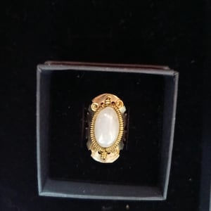Puede incluir: Un anillo dorado con una piedra ovalada blanquecina engastada en un marco ornamentado. El anillo se exhibe en una pequeña caja oscura, mostrando su intrincado diseño y artesanía. El engaste del anillo presenta delicados detalles de filigrana.