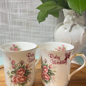 Peut inclure: Deux tasses en porcelaine blanche ornées d'un motif floral de roses roses et de feuilles vertes. Les tasses ont des bords et des anses dorés. Le texte "Ruby Wedding" est imprimé en rouge sur le devant, accompagné d'une petite icône de cloche.