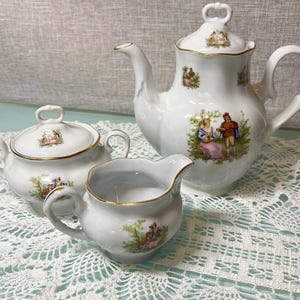 May include: A white porcelain tea set with gold trim, including a teapot, sugar bowl, and creamer. Each item is adorned with a pastoral scene in muted colours. The set is presented on a white lace tablecloth.