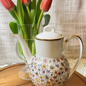 May include: A white porcelain teapot with a floral pattern and gold trim. The teapot is on a wooden tray, and a vase of red tulips is in the background. The teapot has a lid and a curved handle.