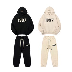 Pu&ograve; includere: Due completi coordinati di abbigliamento casual. Uno &egrave; nero, l'altro color crema. Ogni completo include una felpa con cappuccio e pantaloni della tuta, entrambi con il numero "1997" in grassetto, bianco.