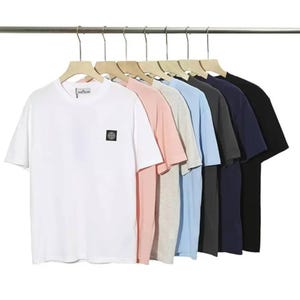 May include: A collection of seven short-sleeved crew neck t-shirts hanging on a metal rack. The shirts are displayed in a row, featuring colours including white, light pink, heather grey, light blue, dark grey, navy blue, and solid black.