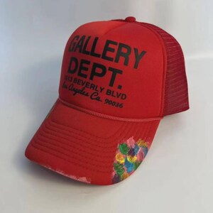 May include: A bright red trucker hat featuring a mesh back and a foam front panel. The front displays black text reading GALLERY DEPT. 7613 BEVERLY BLVD Los Angeles Ca. 90036. The brim has a decorative cluster of colorful paint splatters on the right side.