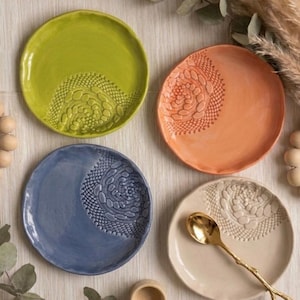 Puede incluir: Cuatro platos de cerámica en tonos verde, naranja, azul y crema, cada uno con un diseño texturizado similar al encaje. Una cuchara dorada descansa sobre el plato crema. Los platos están dispuestos sobre una superficie de tono neutro.