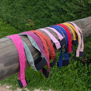 Op de afbeelding: Een verzameling kleurrijke gewichthefbanden in verschillende tinten, waaronder roze, groen, paars, oranje en blauw, wordt getoond op een houten balk. Elke band heeft een zwarte polssteun. De achtergrond toont groen gebladerte.
