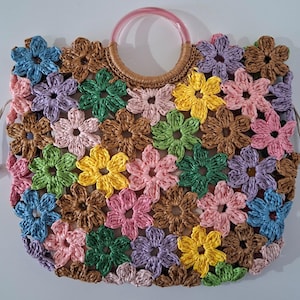 Op de afbeelding: Een geweven draagtas met een bloemenontwerp. De tas heeft een verscheidenheid aan kleurrijke bloemen in tinten blauw, roze, geel, groen en paars. Het heeft een doorzichtige roze handgreep en een geweven bruine handgreepbasis.