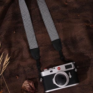 May include: A Leica camera with a silver and black body, featuring a red logo on the front, attached to a black and white gingham patterned camera strap with black leather ends. The camera has a circular lens and a silver top plate.