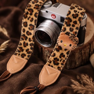 May include: A camera strap featuring a brown and black leopard print pattern with a plush Hello Kitty character attached. The strap includes light tan leather-like end tabs with brown nylon webbing connectors attached to a silver-toned camera body.