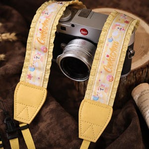 May include: A camera strap featuring a light yellow and white checkered pattern with a ruffled edge. The strap displays the text CookieAnn and cartoon character illustrations. It includes yellow faux leather ends and black plastic attachment clips.