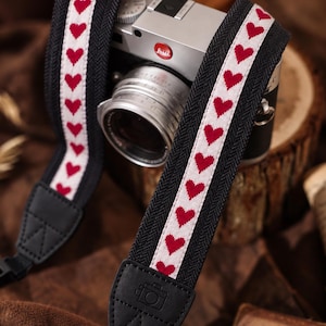 Può includere: Una tracolla per fotocamera con una fascia centrale bianca e un motivo ripetuto di cuori rossi, bordata da tessuto nero a spina di pesce. Include estremità in pelle nera con un'icona di fotocamera in rilievo e fibbie di regolazione nere.