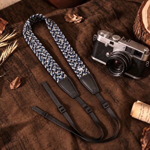 May include: A camera strap featuring a woven blue, white, and black chevron pattern with black leather end tabs and adjustable black nylon webbing straps, positioned next to a silver and black vintage-style camera.