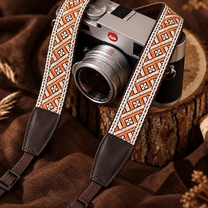 May include: A camera strap featuring a woven geometric pattern in orange, white, and black, attached to dark brown leather end tabs and adjustable brown nylon webbing. The strap is draped over a silver and black Leica camera body.