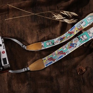 May include: A black and silver camera with a patterned strap featuring comic book illustrations. The strap has tan leather ends. The camera is on a brown surface.