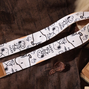 Puede incluir: Correa de cámara blanca con un diseño repetido del personaje de dibujos animados Snoopy en blanco y negro y la palabra "LUCKY". La correa tiene extremos de cuero marrón. El fondo es marrón.