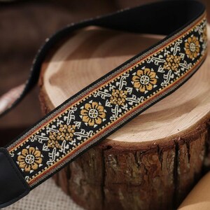 May include: Black camera strap featuring a floral design in gold and white. The strap has a black background with a repeating pattern of flowers and geometric shapes. The strap is made of woven fabric with black leather ends.
