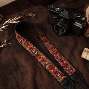 Könnte beinhalten: Ein Kameragurt mit einem lebendigen Blumenmuster aus roten, gelben und grünen Blüten auf beigem Grund, eingefasst in schwarzen Stoff. Der Gurt verfügt über schwarze Lederenden und liegt neben einer schwarzen analogen Kamera.