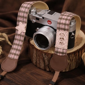 May include: A silver and black vintage-style camera with a brown and pink plaid fabric camera strap. The strap features a light pink star-shaped patch on one side and a rectangular patch with three embroidered cat faces on the other side.