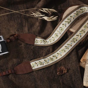 May include: A camera strap with a floral embroidered design in cream, green, and blue. The strap has brown leather ends and is lying on a brown surface next to a camera and a book.