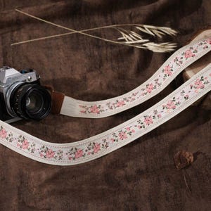 Puede incluir: Una cámara vintage con una correa estampada floral. La correa es de color crema con flores rosas y hojas verdes. La cámara es plateada y negra, con una correa de cuero marrón.