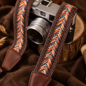 May include: A camera strap featuring a central woven pattern of multicoloured threads in shades of red, orange, yellow, and blue, bordered by solid brown herringbone fabric and finished with brown leather attachment tabs.