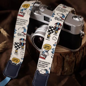May include: A camera strap featuring a cream-colored fabric with a repeating Snoopy cartoon print, hot air balloons, and speech bubbles containing the text "WHERE ARE WE?" and "I'M SPEAKING MY MIND! CHANGES MY OUTLOOK!" with dark blue leather ends.