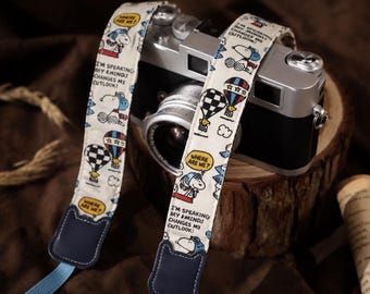 May include: A camera strap featuring a cream-colored fabric with a repeating Snoopy cartoon print, hot air balloons, and speech bubbles containing the text "WHERE ARE WE?" and "I'M SPEAKING MY MIND! CHANGES MY OUTLOOK!" with dark blue leather ends.