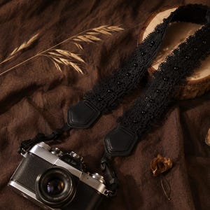 Può includere: Una tracolla per fotocamera nera con intricate rifiniture in pizzo e piccole perline nere, fissata a una fotocamera argento e nera in stile vintage. La tracolla include connettori in similpelle nera che fissano il tessuto al corpo.