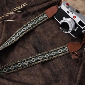 Può includere: Una fotocamera nera e argento con un cinturino in pelle marrone con un motivo geometrico verde, bianco e oro. La fotocamera è appoggiata su una superficie marrone strutturata, accanto a un libro e una fetta di legno.