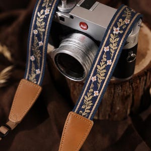 May include: A camera strap featuring a dark blue fabric band with an embroidered pattern of green leaves and small white flowers, accented by tan leather end tabs and adjustable brown nylon webbing straps attached to a silver and black camera.