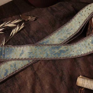 Puede incluir: Una correa de guitarra con un patrón de estrellas azules y doradas. La correa tiene un respaldo y un ribete de cuero marrón. La correa mide aproximadamente 5 cm de ancho.