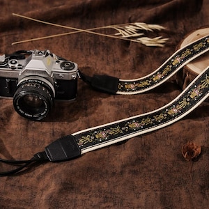 May include: A silver and black Canon camera with a black lens and a decorative camera strap. The strap is black with a white border and a repeating pattern of gold and pink floral designs. The camera and strap are on a brown surface.