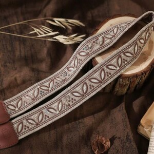 Puede incluir: Correa de cámara con un diseño tejido beige y marrón con un patrón de hojas repetido. La correa tiene extremos de cuero marrón y se muestra sobre una superficie de madera con una estética vintage.