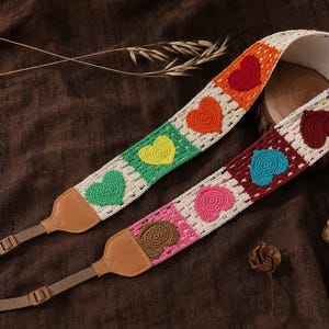 Puede incluir: Una correa para cámara con un patrón de ganchillo de corazones multicolores en rojo, naranja, amarillo, verde, rosa, marrón y azul sobre un fondo crema. La correa incluye extremos de cuero color canela y conectores de nailon ajustables.
