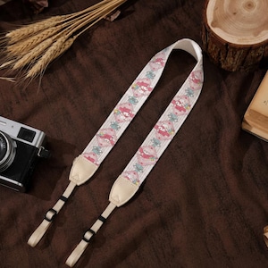 Peut inclure: Une sangle d'appareil photo avec un motif répétitif représentant un personnage à capuche rose avec un nœud rouge, entouré de petites fleurs et de feuilles vertes. La sangle possède des embouts en similicuir beige clair et des boucles réglables.