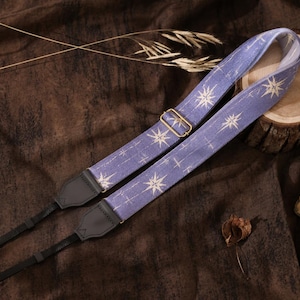 May include: A periwinkle blue camera strap featuring a repeating white starburst pattern. The strap includes a gold-toned metal adjustable slider and black leather-like end tabs with thin black attachment cords for securing to a camera body.