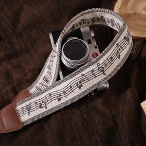 Peut inclure: Une courroie d'appareil photo composée d'une bande en tissu crème ornée d'une portée musicale et de notes noires, bordée d'un motif à chevrons gris. La courroie comprend des embouts en cuir marron avec des coutures et est fixée à un appareil.