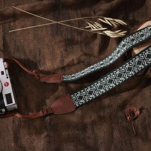 Puede incluir: Una cámara negra y plateada con una correa estampada. La correa presenta un diseño geométrico en blanco y verde oscuro. La cámara está sobre una superficie marrón, con una rodaja de madera y un libro en el fondo.