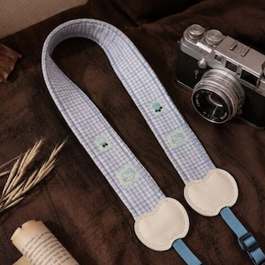 May include: A camera strap featuring a light blue and white gingham pattern with small embroidered character motifs. The strap includes cream-coloured apple-shaped leather tabs at the ends, connected to adjustable blue nylon webbing and plastic buckles.