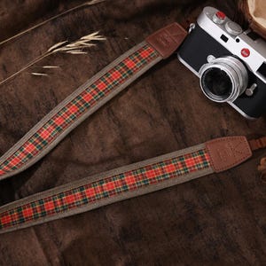 Può includere: Una tracolla per fotocamera con un motivo in tessuto scozzese rosso, verde e giallo, bordi intrecciati color marrone chiaro ed estremità di fissaggio in pelle marrone. La tracolla è collegata a una fotocamera Leica argento e nera con obiettivo.