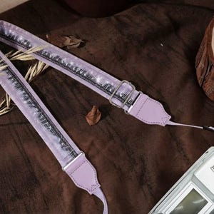 May include: A lavender camera strap featuring a repeating pattern of white musical notes and staves on a dark background. The strap includes a silver metal adjustable buckle and light purple leather-like end tabs attached to a white camera body.