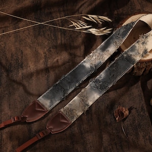Puede incluir: Una correa para cámara con un diseño de tela desgastada y deshilachada en tonos gris carbón y blanco roto. La correa incluye extremos de cuero marrón oscuro y conectores ajustables de nailon marrón para fijarla al cuerpo de la cámara.