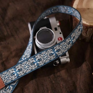Puede incluir: Una correa de cámara con estampado azul y blanco con un extremo de cuero marrón está colocada sobre una cámara plateada. La correa presenta un diseño floral repetido. La lente de la cámara es visible. El fondo incluye una superficie de madera y un libro.