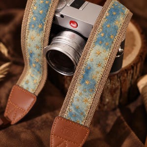 May include: A camera strap featuring a blue fabric centre with gold embroidered star patterns, bordered by brown herringbone trim and finished with brown leather end tabs embossed with a small camera icon, attached to a silver and black camera.