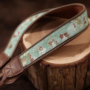 Puede incluir: Una correa de cámara con un estampado floral en tonos verdes, rojos y blancos. La correa tiene extremos de cuero marrón y un borde de espiga marrón. Descansa sobre una superficie de madera.