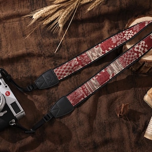 Può includere: Una fotocamera digitale nera con piastra superiore argentata fissata a una tracolla in tessuto. La tracolla presenta un motivo ripetuto di forme geometriche e floreali rosse, bianche e grigie, con estremità in pelle nera e fibbie regolabili.