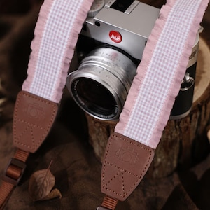 May include: A camera strap featuring a light pink and white gingham fabric centre with ruffled edges, attached to brown leather end tabs with embossed camera icons and adjustable brown nylon webbing straps, connected to a silver and black camera.