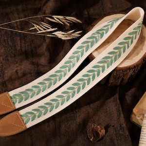 May include: A camera strap featuring a cream-colored fabric band embroidered with a repeating green leaf pattern. The strap includes tan leather end tabs with visible stitching and black plastic hardware connectors at both ends of the strap.