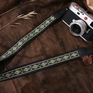 Puede incluir: Correa de cámara negra con un patrón floral en verde y dorado. La correa está sobre una superficie marrón junto a una cámara negra y plateada. La correa tiene un respaldo negro y está diseñada para una cámara.