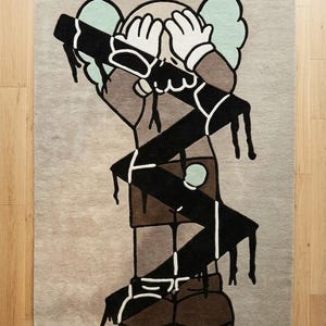 Overhead view of abstract designer rug featuring bold character artwork, contemporary statement carpet in neutral beige, black, and brown tones.