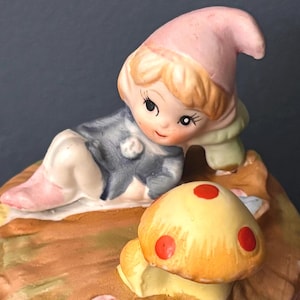 May include: A ceramic figurine of a small elf with blonde hair wearing a pink hat and a blue tunic, reclining on a brown surface next to a yellow mushroom with red spots. The elf has large eyes and is resting its head on a green pillow-like base.